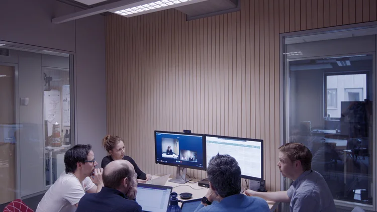Group of five people in a conference room participating in a video call projected on two monitors, with laptops and documents on the table.