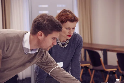 Two people, a man and a woman, leaning over a table and looking down at something, in a room with beige curtains and chairs in the background.