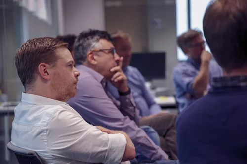 A group of men sitting attentively during a meeting or presentation at an office. Some are focused, others pondering, with a window and office equipment visible in the background.